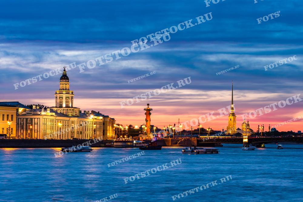 Фотография показывает классический вид Санкт-Петербурга в сумерках: Кунсткамеру, Ростральные колонны и Петропавловскую крепость с золотым шпилем. Городской пейзаж купается в мягком свете заходящего солнца, который рисует на небе градиент от тёплых оранжево-розовых до глубоких синих оттенков, отражающихся в водах Невы. Изображение подходит для статей о культурных памятниках, туристических проспектах и в качестве изображения для любителей вечернего городского ландшафта.